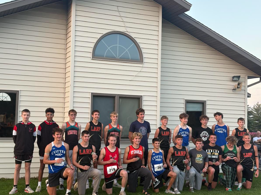 A group of male cross-country runners pose for a team photo outside a building, wearing jerseys from various schools, including LARP, Cotter, and St. Charles. Some runners kneel in the front row while others stand behind them, several holding medals or race items.