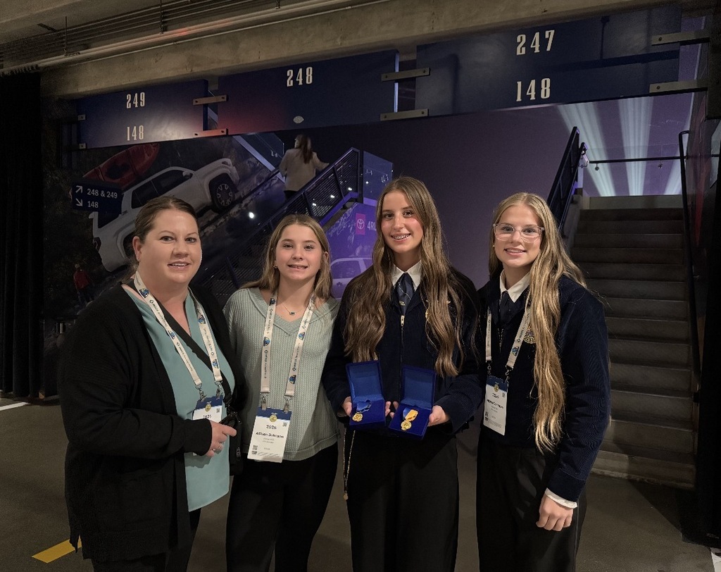 Four women pose for a photo at an event venue. Two wear navy-blue formal uniforms with gold details, while the others wear casual tops and lanyards with event badges. One of the uniformed women holds two open blue cases displaying gold medals. A staircase and event signage are visible in the background.