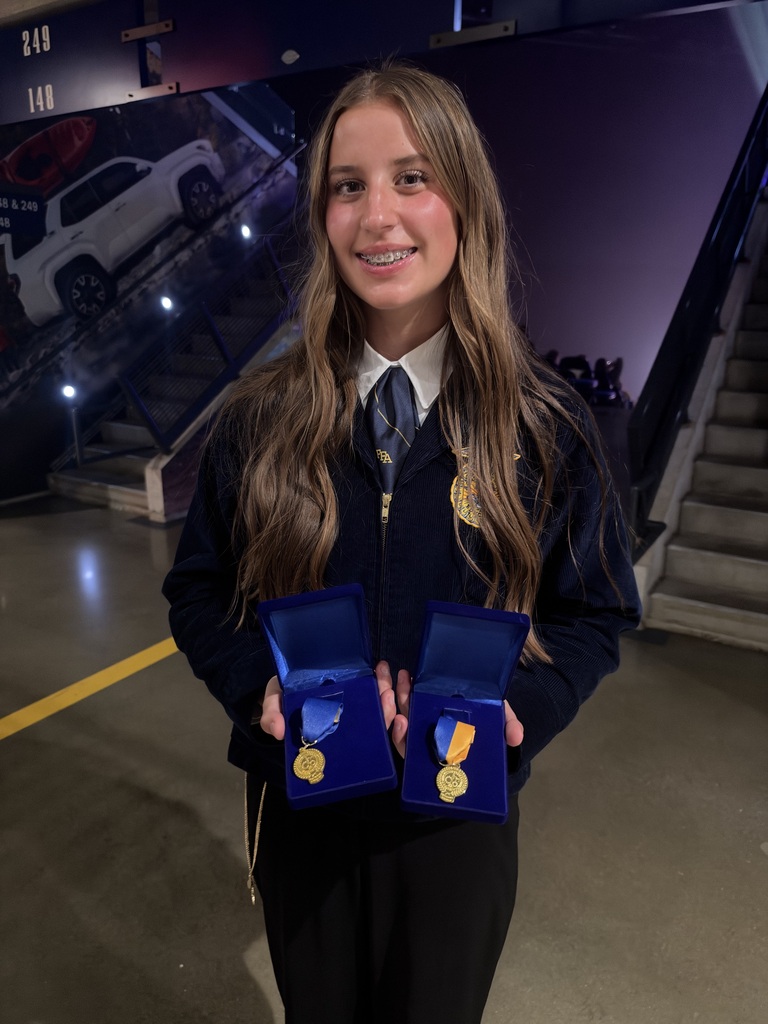 A young woman in a navy-blue formal uniform with gold details smiles while holding two open blue medal cases, each displaying a gold medal. She poses in front of a staircase inside an event venue.