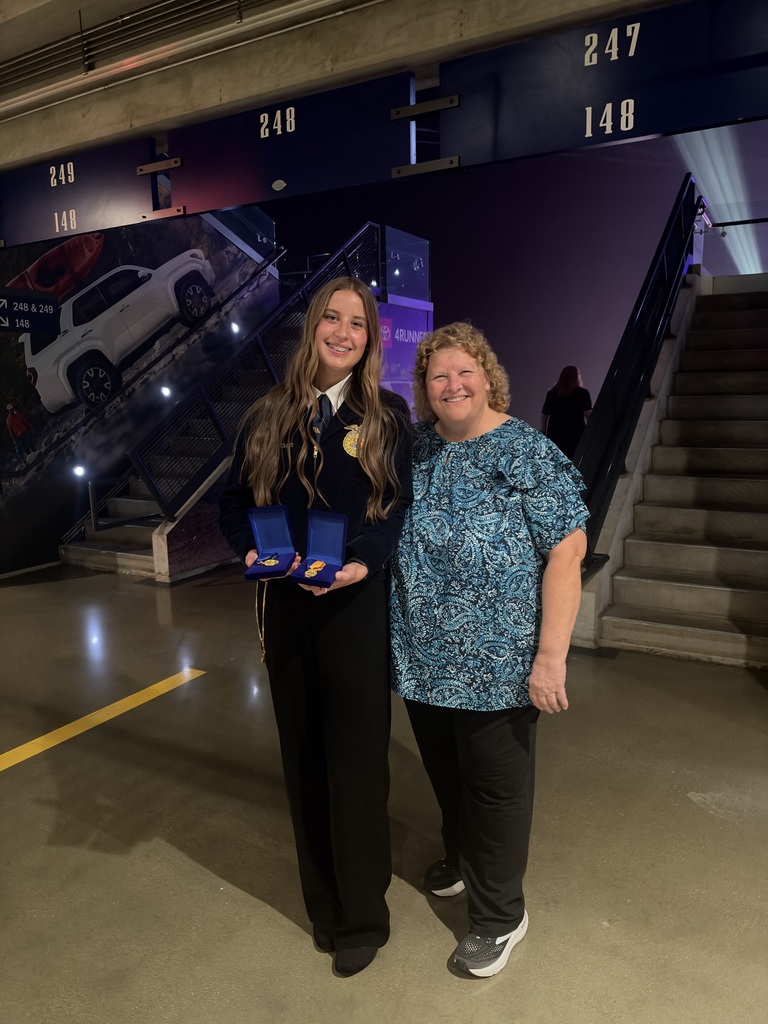 A young woman in a dark formal uniform with a gold emblem stands beside a smiling woman in a blue patterned shirt. The young woman holds two open blue medal cases displaying gold medals. They pose for a photo near a staircase inside an event venue.