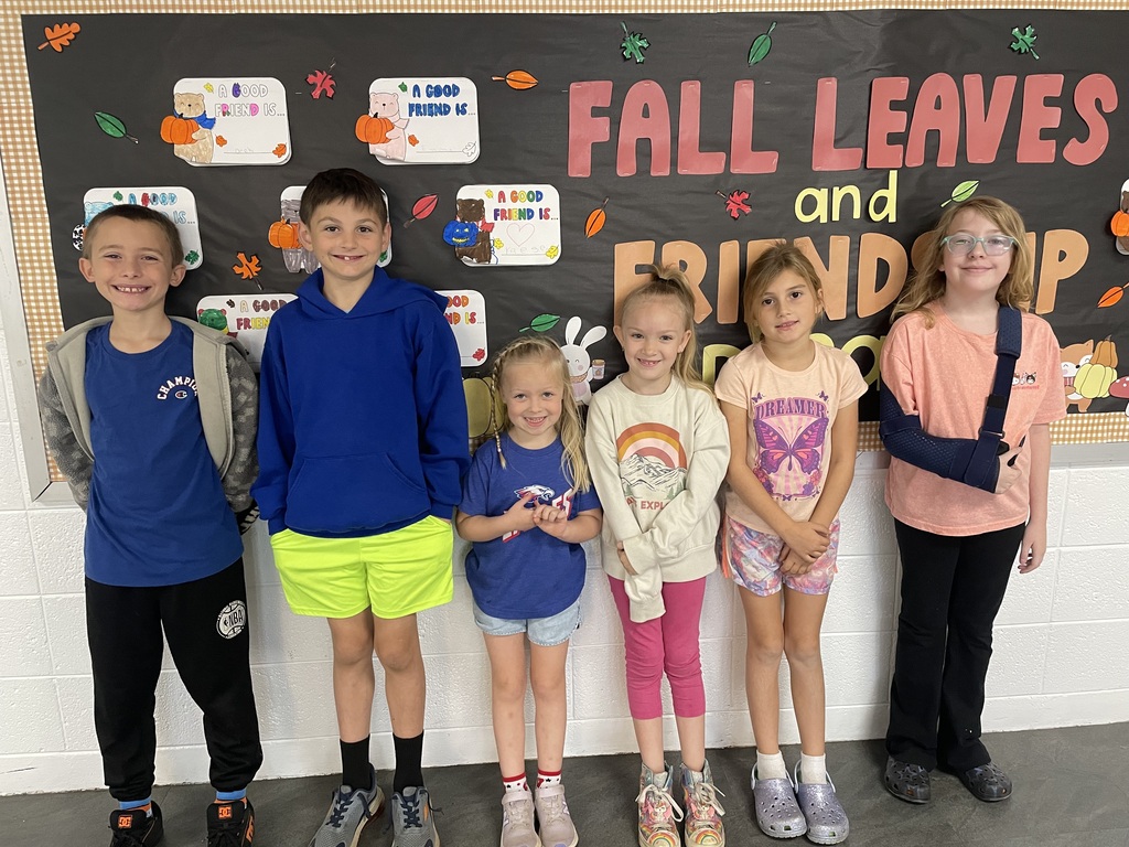 Six children pose for a photo in front of a classroom bulletin board decorated with fall leaves and the words “Fall Leaves and Friendship.” The children wear colorful casual clothing, and the child on the far right has their arm in a sling. Small artwork with the theme “A Good Friend Is…” decorates the bulletin board.