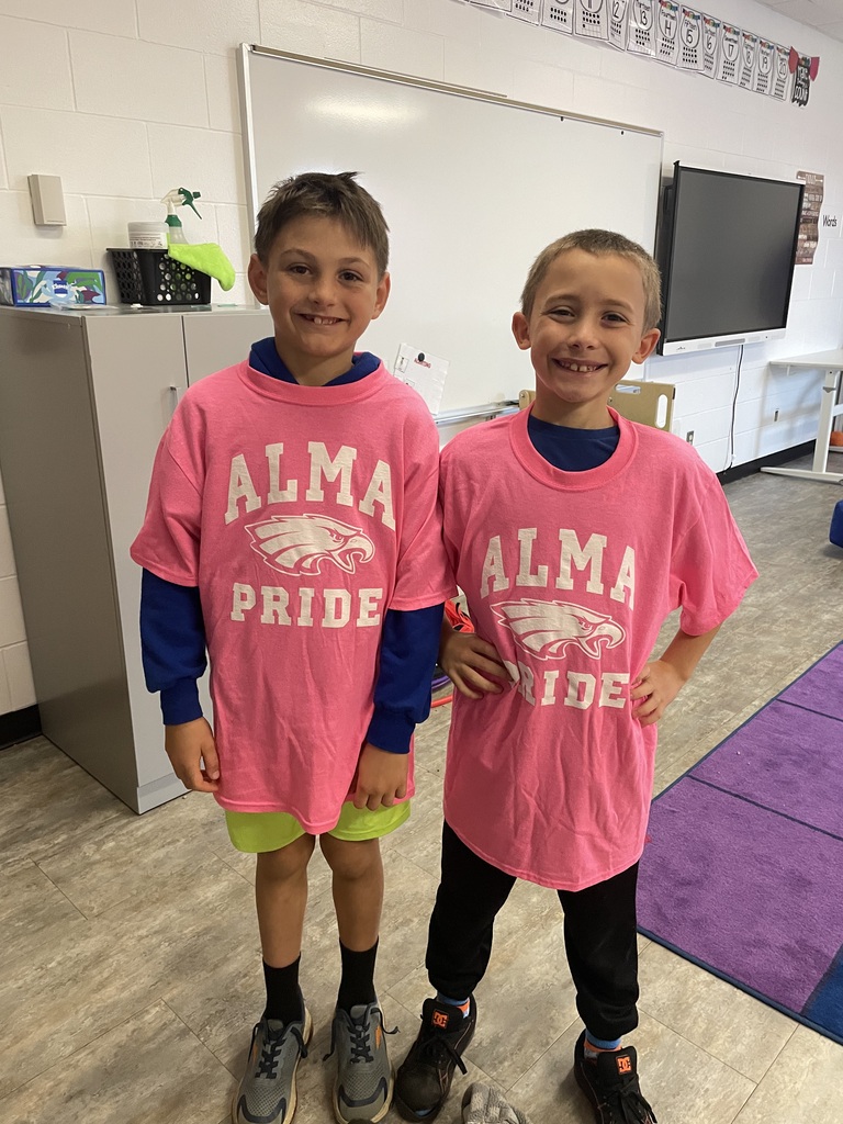 Two students smile for a photo in a classroom while wearing matching pink “Alma Pride” T-shirts featuring an eagle logo. One wears neon yellow shorts and gray sneakers, while the other wears black pants and black sneakers. A whiteboard, cabinets, and a purple classroom rug are visible in the background.