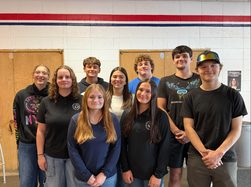 Nine students pose for a group photo in a school hallway, standing in two rows and smiling at the camera. They wear casual clothing, mostly dark shirts, with one student wearing a blue shirt and another wearing a white top. Red and blue stripes run along the white brick wall behind them, with wooden doors on either side.