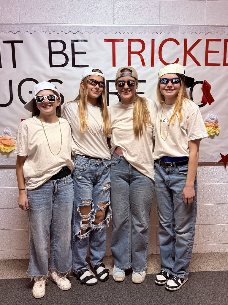 Four students pose together in matching white T-shirts, baggy jeans, backward baseball caps, sunglasses, and gold-colored chains for a group costume. They stand in front of a school bulletin board with large red and black lettering, colorful decorations, and a red ribbon.
