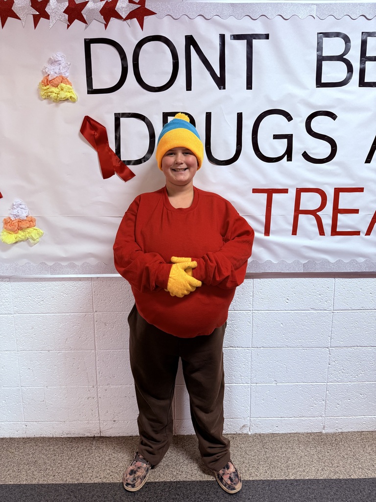 A student smiles for a photo wearing a red sweatshirt, brown pants, yellow gloves, and a blue-and-yellow knit hat as a costume. They stand in front of a school bulletin board decorated with red stars, ribbons.