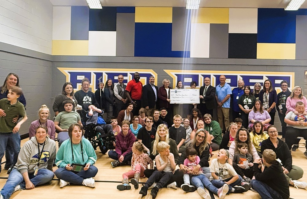 In a vibrant gymnasium, a large group of individuals is gathered, capturing a moment of community and collaboration. The photograph features representatives from the AMA ESD Board of Education, Pied Piper School Students and Staff, representatives from Blue Horizons, and representatives from the Community Foundation for Northeast Michigan.  At the center, several adults are holding a large check, symbolizing a significant contribution or partnership. The students, interspersed among the adults, add a youthful energy to the scene, some smiling brightly at the camera. The Pied Piper School staff, easily identifiable by their supportive presence, are seen engaging with the students and other representatives. The Blue Horizons and Community Foundation for Northeast Michigan representatives stand alongside, their participation highlighting the broad community support for the initiative.  The background of the gymnasium features colorful wall panels in shades of blue, yellow, and gray, adding a dynamic backdrop to the gathering. The overall atmosphere is one of warmth, cooperation, and shared purpose, reflecting a successful collaboration between various educational and community organizations.
