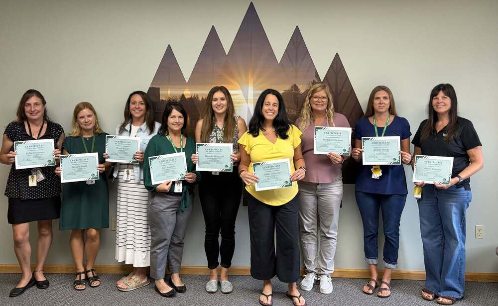 Nine women stand in a line indoors holding framed Certificates of Appreciation in front of a wall decal featuring a mountain silhouette graphic.