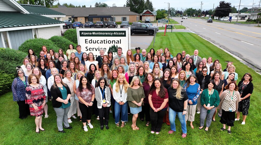 A large group of staff members posing together outdoors on the grass in front of the Alpena-Montmorency-Alcona Educational Service District sign.