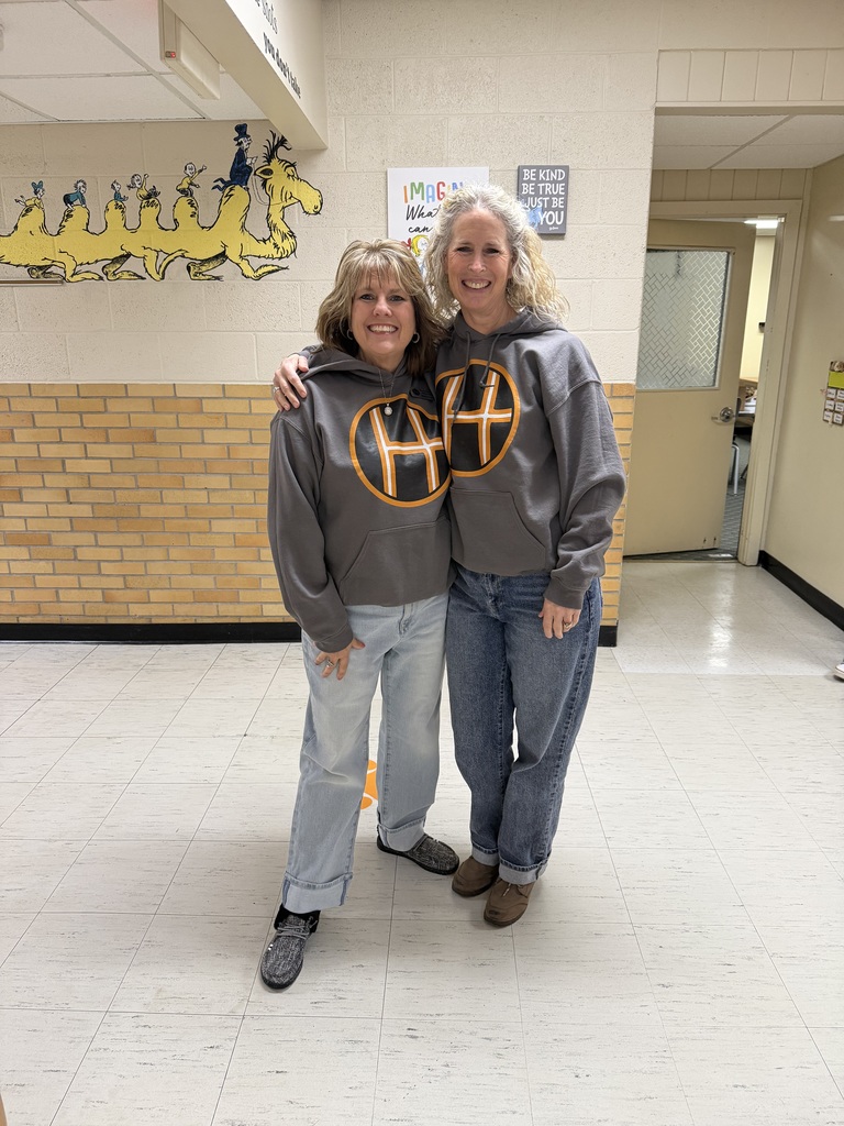 A full-length photo of two smiling women standing side-by-side with their arms around each other in a school hallway. They are wearing matching dark grey hoodies featuring a large logo of a stylized black "H" inside an orange circle. The woman on the left has shoulder-length light brown hair and wears light-wash cuffed jeans with grey sneakers. The woman on the right has long, wavy blonde hair and wears dark-wash jeans with brown shoes. Behind them is a yellow brick wall decorated with a Dr. Seuss mural of yellow creatures and inspirational posters, one reading "Be Kind, Be True, Just Be You."