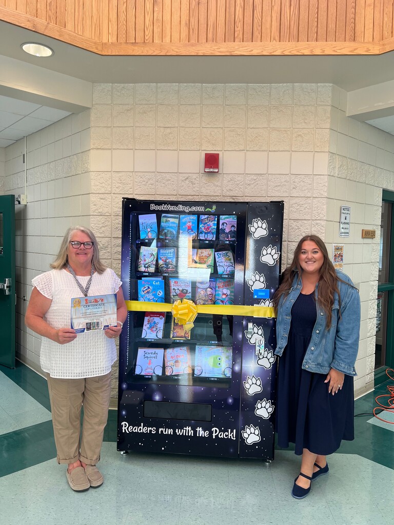 Two women stand beside a newly installed children’s book vending machine in a school hallway. The vending machine has a galaxy-inspired design with colorful northern-lights effects, white paw prints, and the words “Readers run with the Pack!” across the bottom. The machine is filled with colorful children’s books and is decorated with a yellow ribbon for its ribbon-cutting ceremony. The woman on the left holds a certificate celebrating the new vending machine, while the woman on the right smiles beside the machine.