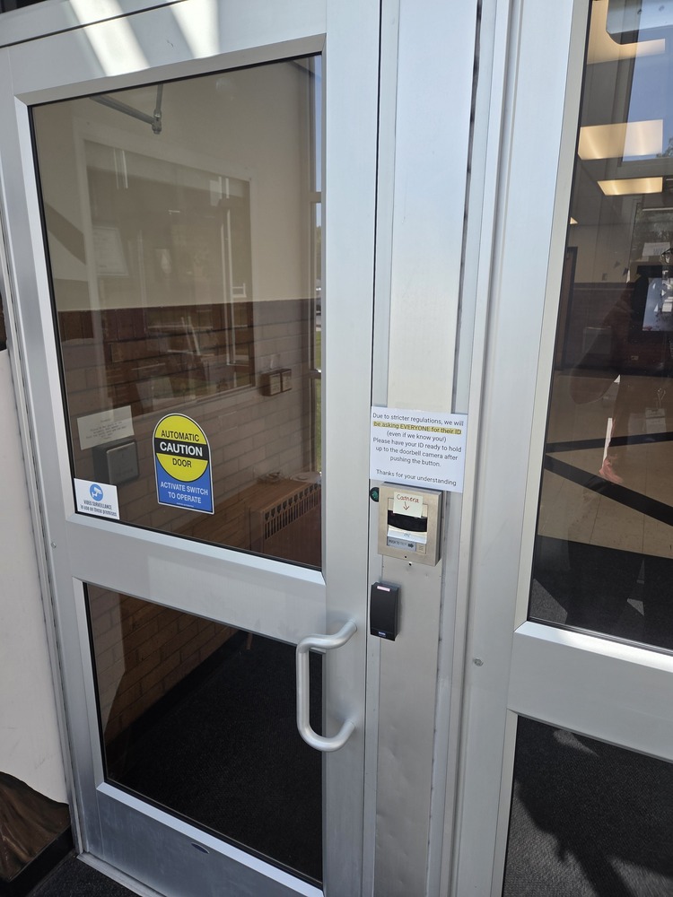 The front door of Lehi Elementary. A camera and sign are posted about visitors needs to check in with ID, there is another post about recording being in progress.