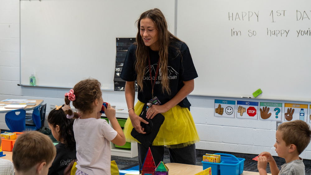 Teacher standing in front of kindergarten class. Whiteboard behind reads "happy 1st day"