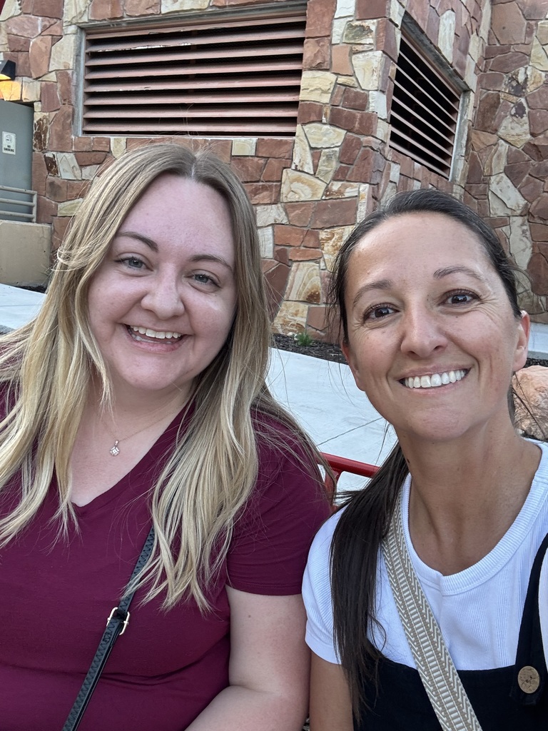 Two women smiling, one with blond hair and a red shirt, the other with dark brown hair, a white shirt and black overalls