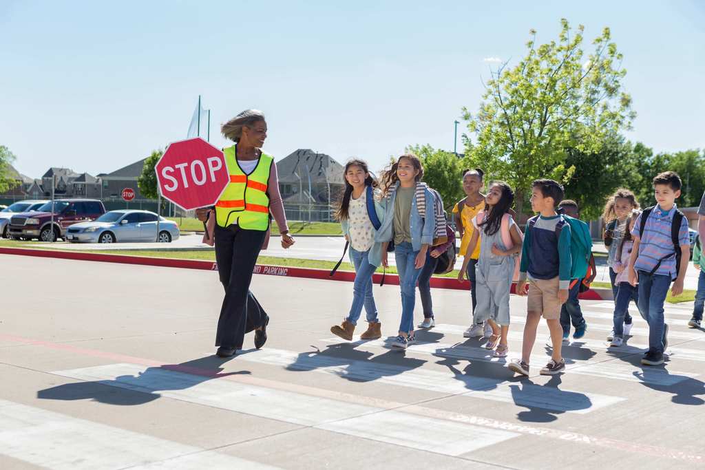 A female crossing guard holding a stop sign helps nine children cross a street.
