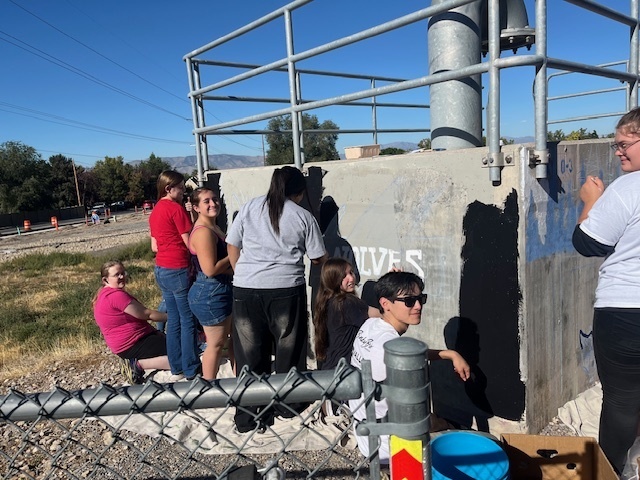 Alt text: A group of high school students works on painting the side of a concrete wall outdoors. Some students stand while others sit on a tarp, filling in the background with black paint around the word “T-WOLVES.” The wall has a metal railing structure on top, and the group appears to be collaborating and smiling. A chain-link fence and painting supplies are visible in the foreground, with trees and mountains in the background under a clear blue sky.