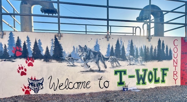 Alt text: A mural painted on a concrete wall shows a wolf pack running through a forest of evergreen trees. Red paw prints lead toward the center of the mural. The words “Welcome to T-WOLF COUNTRY” are painted in a combination of cursive black lettering and large green block letters outlined in blue. A fierce wolf head logo is on the left side, and a red vertical “COUNTRY” is on the right edge of the mural. The background includes railings and utility pipes above the wall, with sunlight shining through.