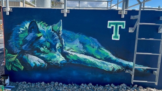 Alt text: A mural on a blue wall depicts a large wolf painted in cool blue and green tones, lying down with its head resting on its front paws and eyes looking forward. A small green leaf is painted near its snout. To the right, a bold green and white letter “T” is displayed. A metal ladder is mounted on the wall above the wolf, and gravel covers the ground below.