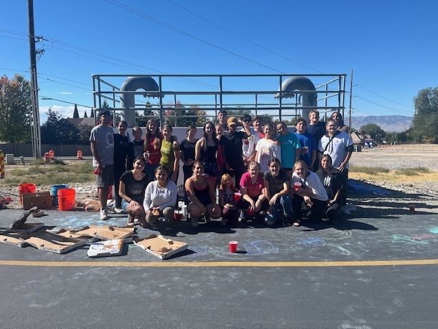 Alt text: A large group of high school students poses together in front of a concrete wall and metal railing structure on a sunny day. The students are smiling, some holding paint supplies, wearing casual clothes after working on a mural project. Pizza boxes and painting materials are scattered on the pavement in front of them, and chalk outlines are visible on the ground. Trees, power lines, and mountains can be seen in the background under a clear blue sky.