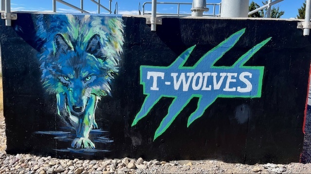 Alt text: A mural on a black wall features a large, detailed painting of a wolf with glowing blue and green tones, crouched low as if stalking forward with piercing eyes. To the right of the wolf, bold block letters read “T-WOLVES” in white with a blue and green outline. The text is set against a stylized claw mark graphic with three slashes. Metal railings and pipes are visible at the top of the wall, and gravel lines the ground below.