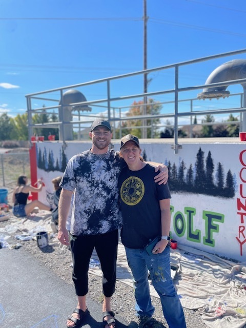 Alt text: Two adults stand side by side smiling with arms around each other in front of a mural that reads “T-WOLF COUNTRY.” The mural features a forest of black trees and green outlined letters. Paint supplies and tarps are scattered on the ground, and students can be seen working on the mural in the background. The sky is clear and blue, and metal railings and pipes run above the wall.
