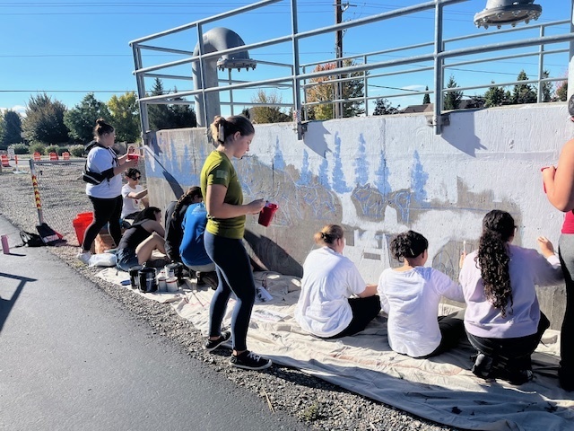 Alt text: A group of high school students sits and stands along a large concrete wall, painting the base layer of a mural on a sunny day. Several students are wearing casual clothes and holding red cups with paint, working on outlining trees and letters. A tarp covers the ground, and paint cans are scattered nearby. Metal railings and industrial structures are above the wall, with trees and traffic cones visible in the background.