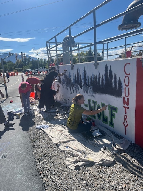 Alt text: Several students work together to paint a large mural on an outdoor concrete wall near a roadway. One student in a yellow shirt sits on a tarp painting the green “T-WOLF” letters, while others add details to the surrounding forest and wolf imagery. Traffic cones and construction barriers line the street in the background under a bright blue sky. Metal railings and pipes run along the top of the wall.