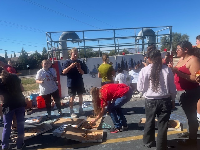Alt text: A group of high school students gathers in front of a mural wall during a sunny day, eating pizza after working on the project. Several open pizza boxes are spread on the pavement, and some students are standing or sitting while talking. The mural in the background features forest silhouettes and the words “Welcome to T-WOLF Country.” Metal railings and pipes run along the top of the wall, and chalk drawings are visible on the ground.