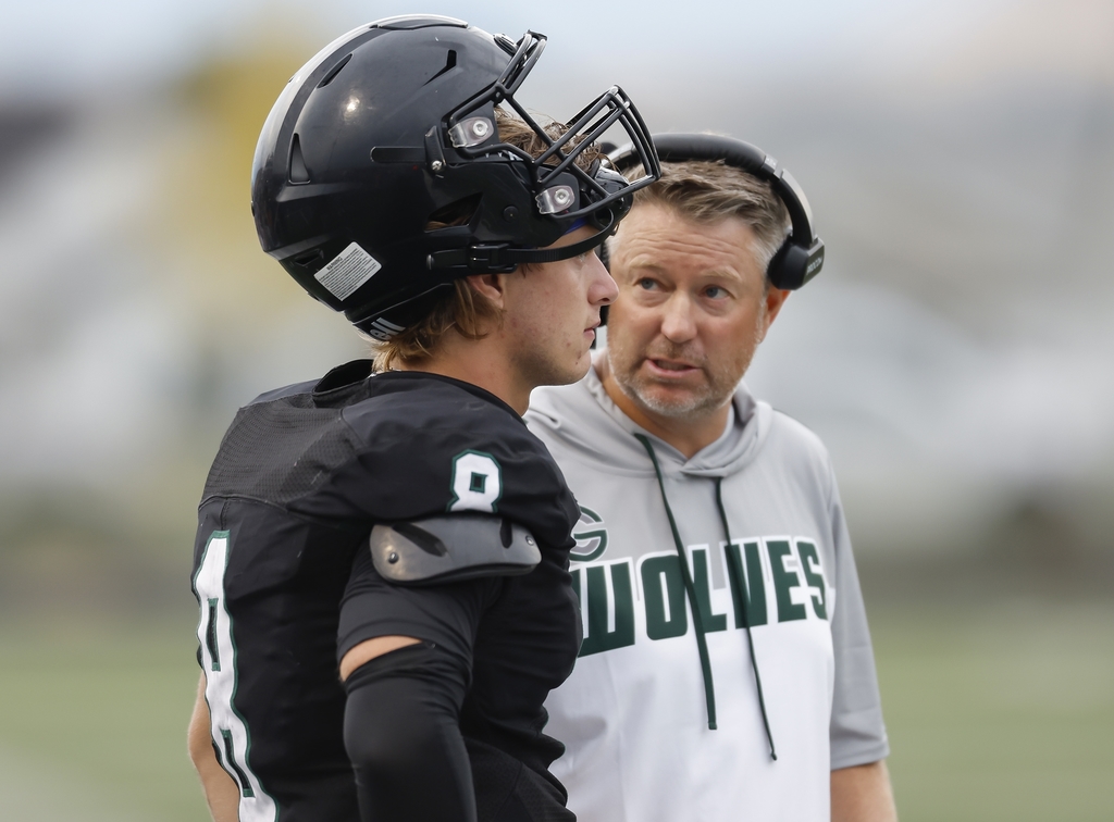 A football coach wearing a headset speaks closely with a player in a black football uniform and helmet during practice or a game. The coach appears focused and engaged, offering instruction while the player listens attentively on the field.