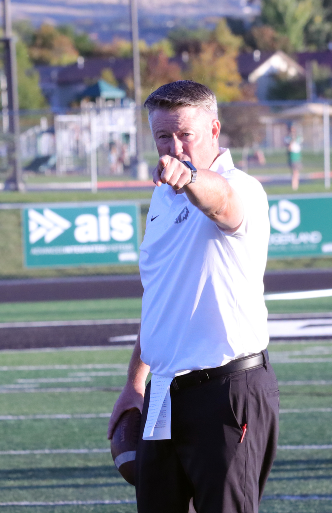 A football coach stands on a turf field wearing a white polo and black pants, holding a football and pointing directly toward the camera. Stadium signage and trees are visible in the background, suggesting an active game or practice setting.