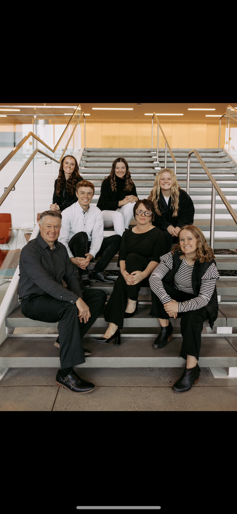 A family of eight poses together on a wide indoor staircase. The group is dressed in coordinated black, white, and neutral clothing, smiling toward the camera in a well-lit, modern building.