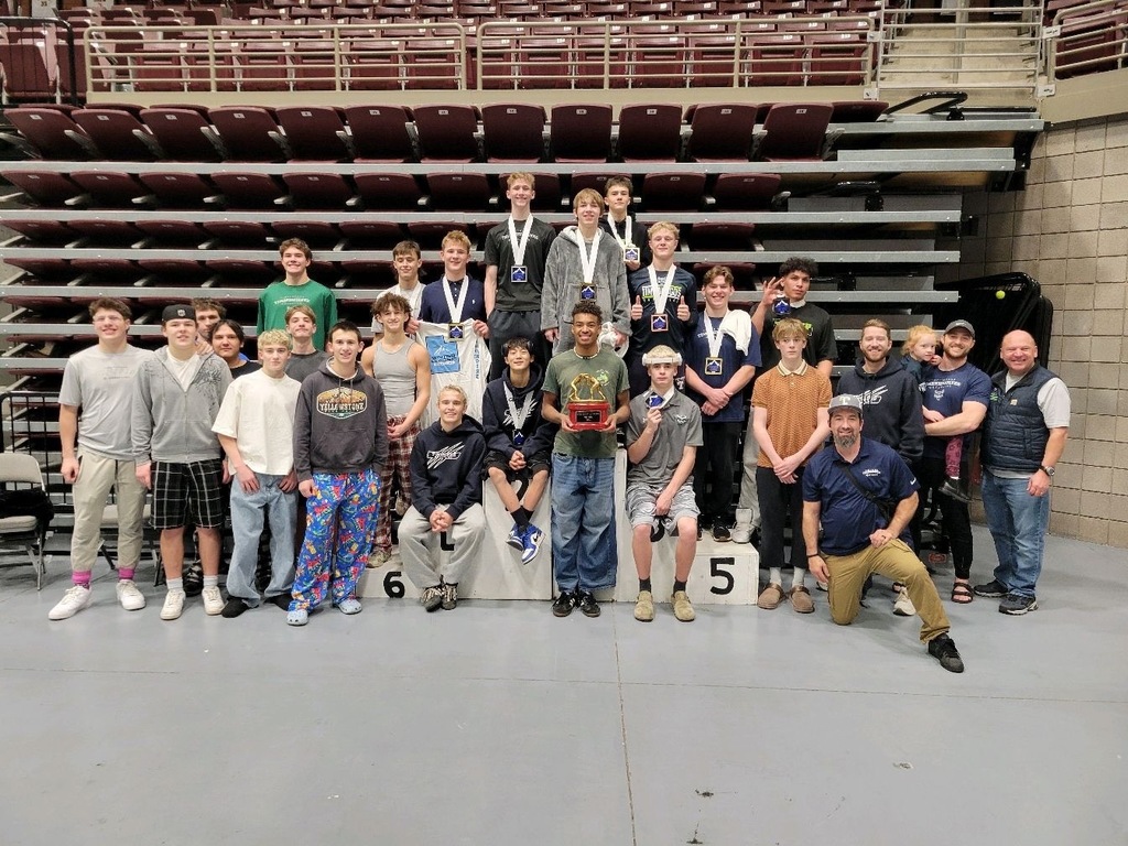 A large group of high school wrestlers and coaches pose inside an indoor arena after a tournament. Several wrestlers stand on a podium wearing medals, while teammates and coaches stand around them smiling. One wrestler in the center holds a first-place team trophy. Empty maroon stadium seats fill the background, highlighting the post-tournament setting.