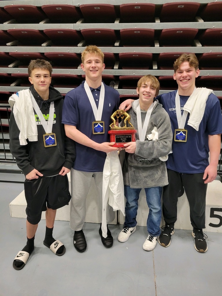 Four high school wrestlers stand shoulder to shoulder on a podium wearing medals. The two wrestlers in the middle hold a first-place team trophy together. All four are smiling, dressed in athletic clothing, with folded wrestling mats and empty maroon arena seats visible behind them.