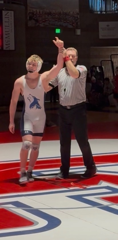 “A referee raising the hand of a Timpanogos wrestler in a white singlet with a wolf logo after winning a match.”