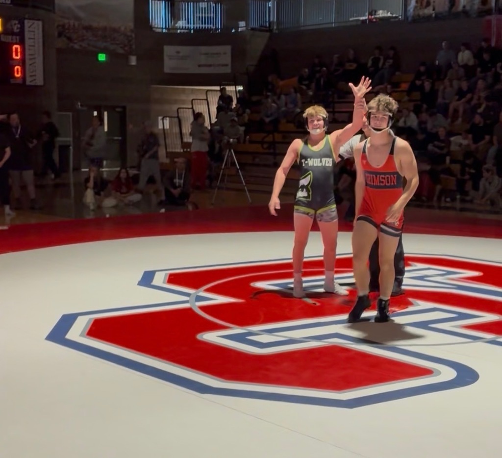 “A referee holding up the arm of a Timpanogos wrestler in a navy T-Wolves singlet as the crowd watches from the stands.”