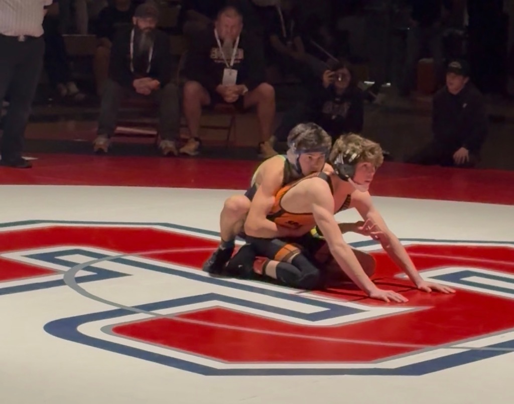 A Timpanogos wrestler riding his opponent from behind on the center mat during a match, with spectators seated at mat-side.”