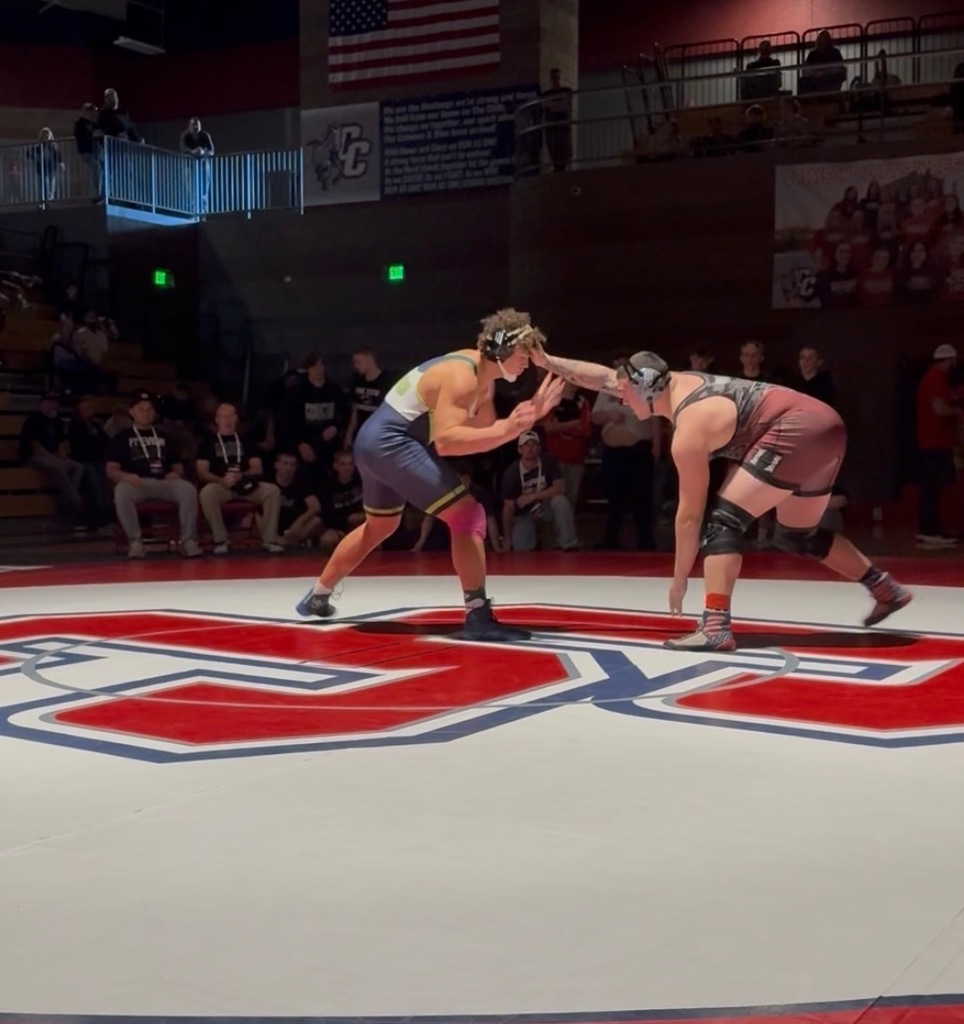 Two high school wrestlers crouched and locked up at the start of a match on a red, white, and blue wrestling mat in a crowded gym.”