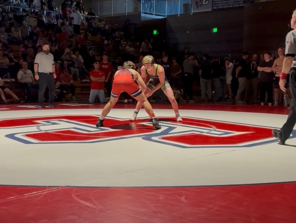 Two wrestlers standing chest-to-chest in a neutral tie position at center mat, preparing to engage as a referee watches nearby.”