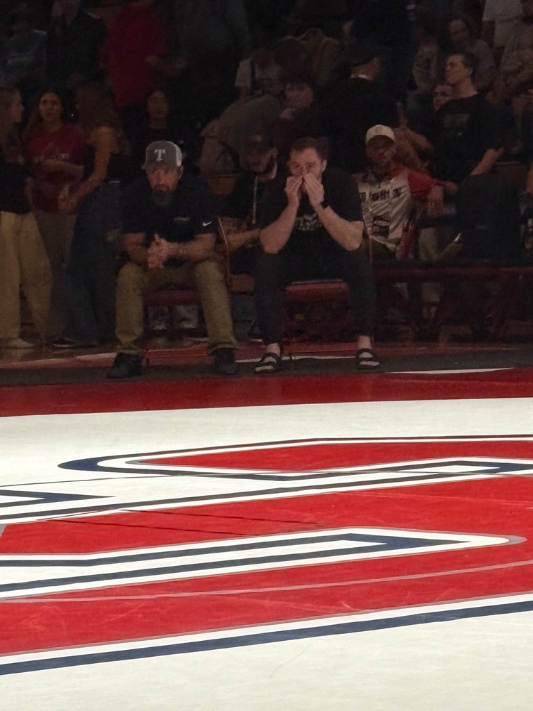 Two Timpanogos coaches sitting on the edge of the mat during a match, watching intently as the crowd sits behind them.”