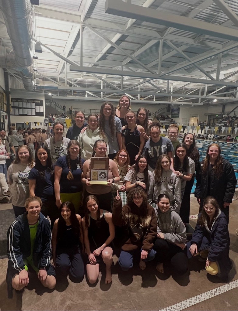 Timpanogos High School girls swim team posing together on a pool deck with a large indoor competition pool behind them; one athlete is holding a championship plaque at the center of the group.”