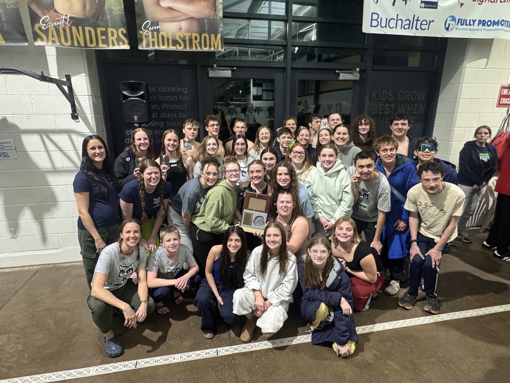 Timpanogos swimmers and coaches kneeling and standing together in front of a glass entrance at a natatorium, with one athlete holding a Region Championship plaque in the middle.”