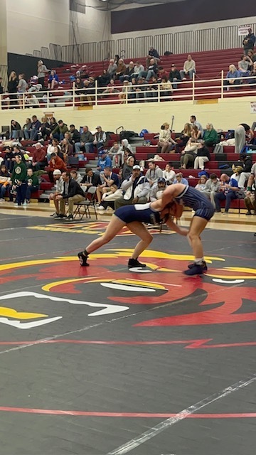 “Two high school wrestlers in a neutral tie position in the center of a competition mat, competing in front of bleachers filled with spectators.”