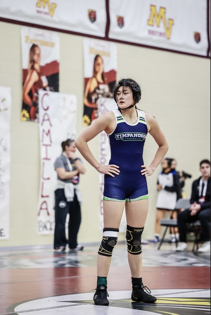 Timpanogos wrestler Liv Ahn standing at the center of a wrestling mat in a navy T-Wolves singlet with her hands on her hips, wearing headgear and knee pads while officials and coaches look on.”