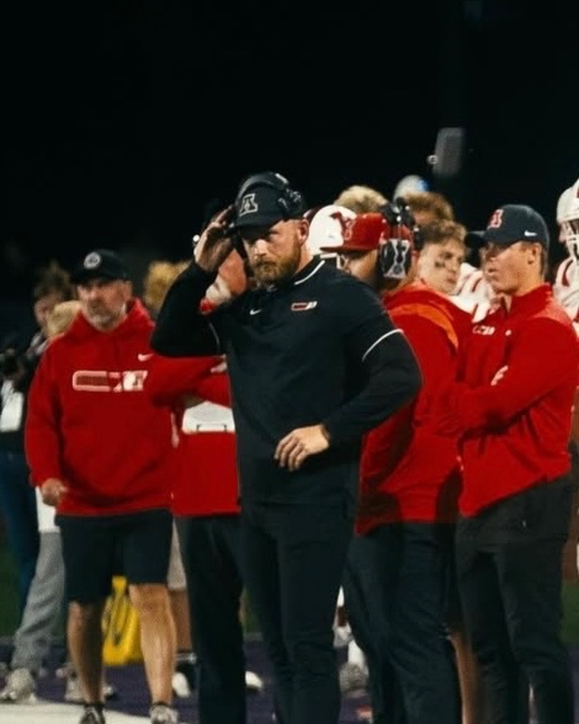A football coach in black athletic gear and headset stands on the sideline at night while players and staff in red stand behind him.