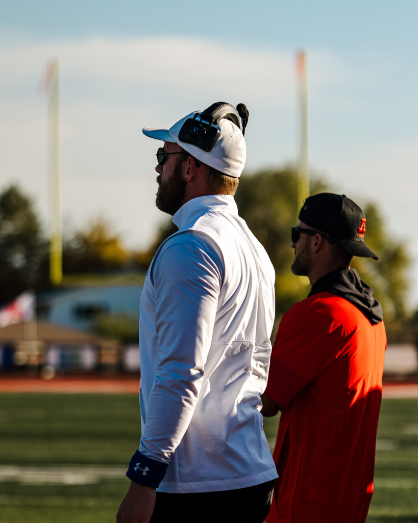 Two football coaches stand on a field during daytime, viewed from the side, both wearing caps and athletic gear while watching the action on the field.