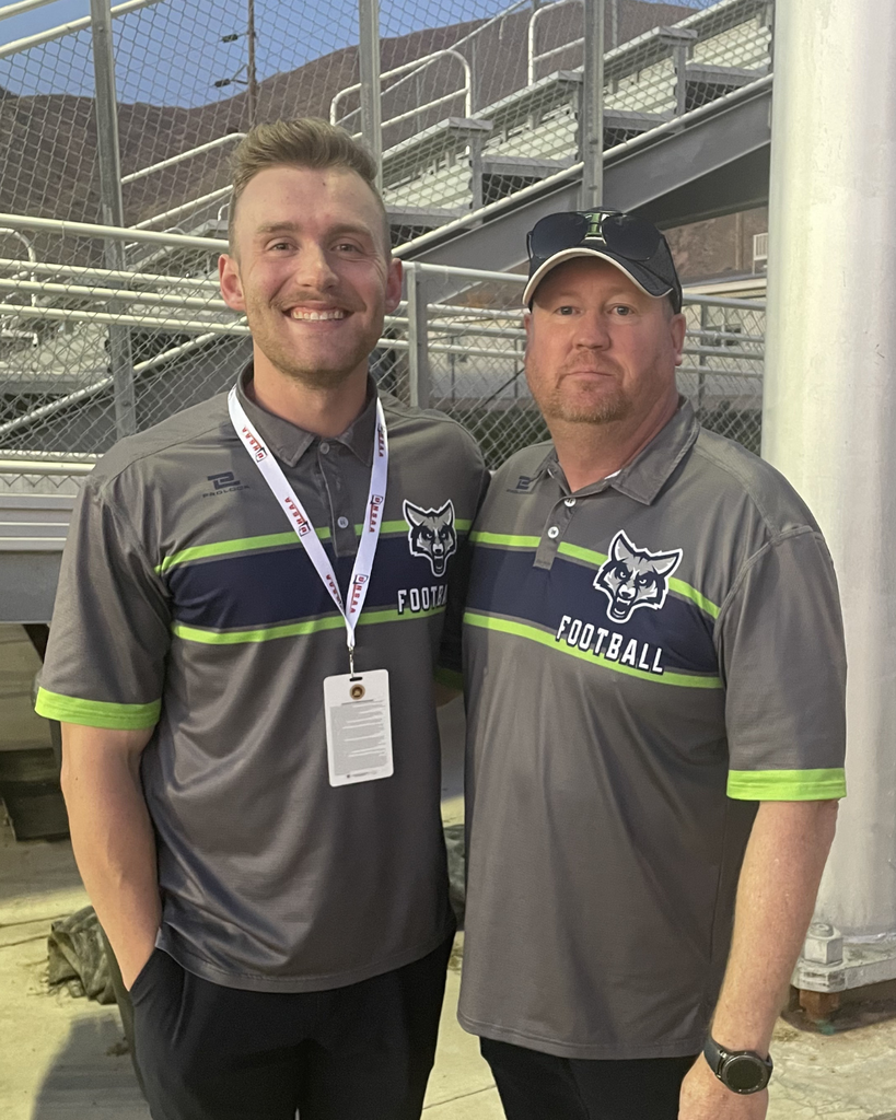 Two football staff members wearing matching gray team polos pose together and smile in front of stadium bleachers.