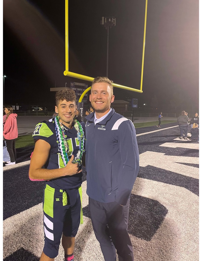 A football player wearing a navy and neon green uniform and celebratory leis stands with a coach on the field at night under the goalpost, both smiling at the camera.