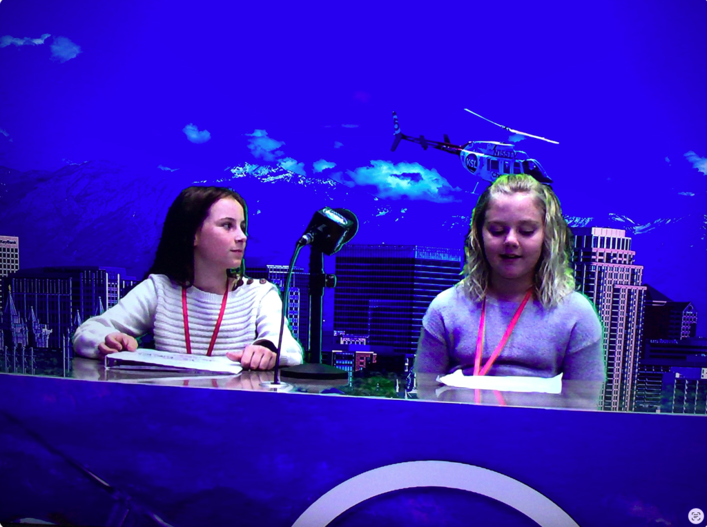 Two students sitting down with a microphone sitting on the desk between them. The background is the city. They are doing a news report.