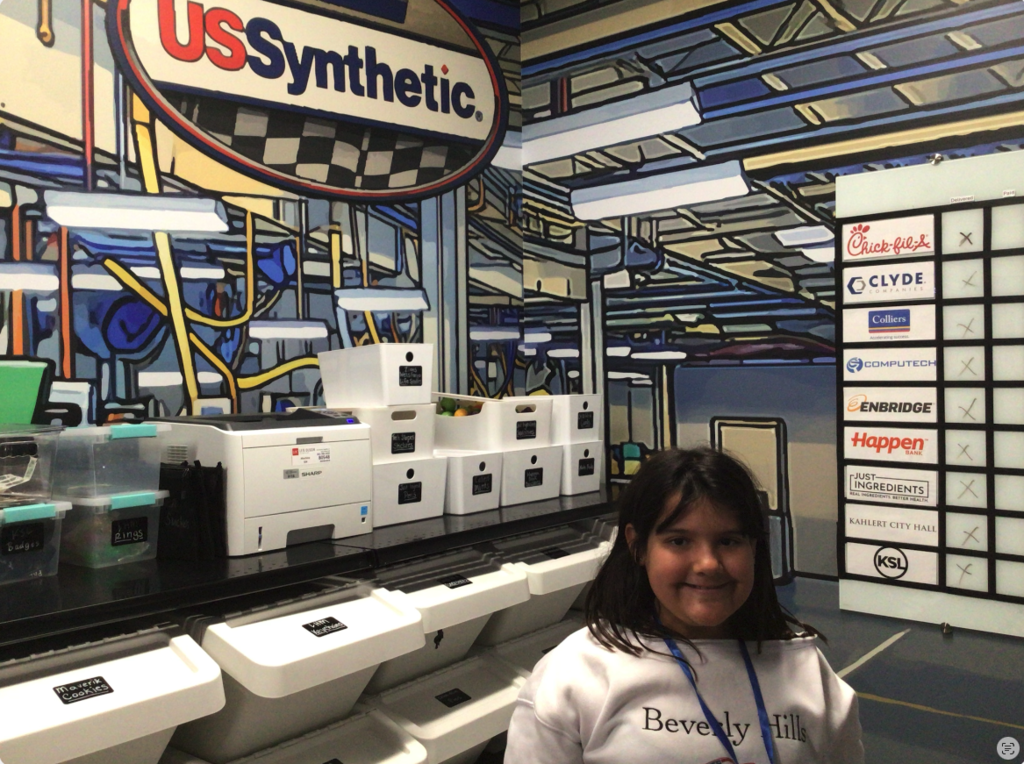 Student smiling standing in her station learning about the business "USSynthetic". In the background there is a printer and some containers.