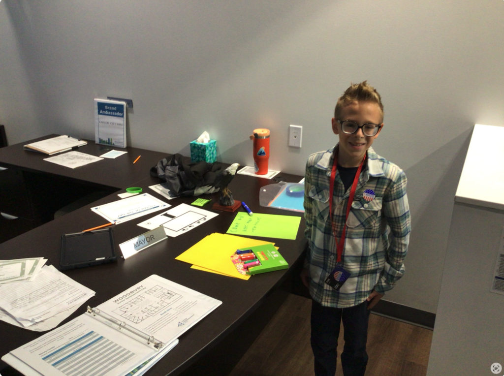 Student standing smiling to the camera, in front of the desk. There is a desk sign saying "Mayor". There is a binder on the desk, the student also has a pin saying "Voted" on his shirt.