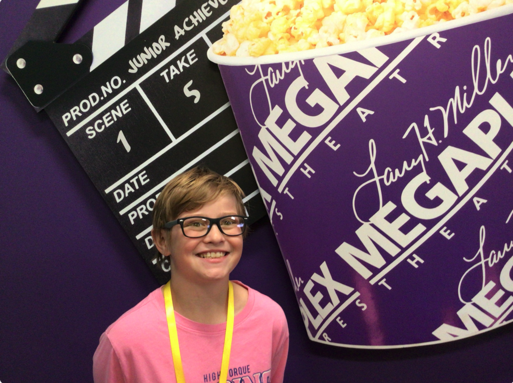 Student smiling in front of a "Megaplex Theater" sign. The sign is a popcorn purple bucket, behind the bucket there is a film slate.
